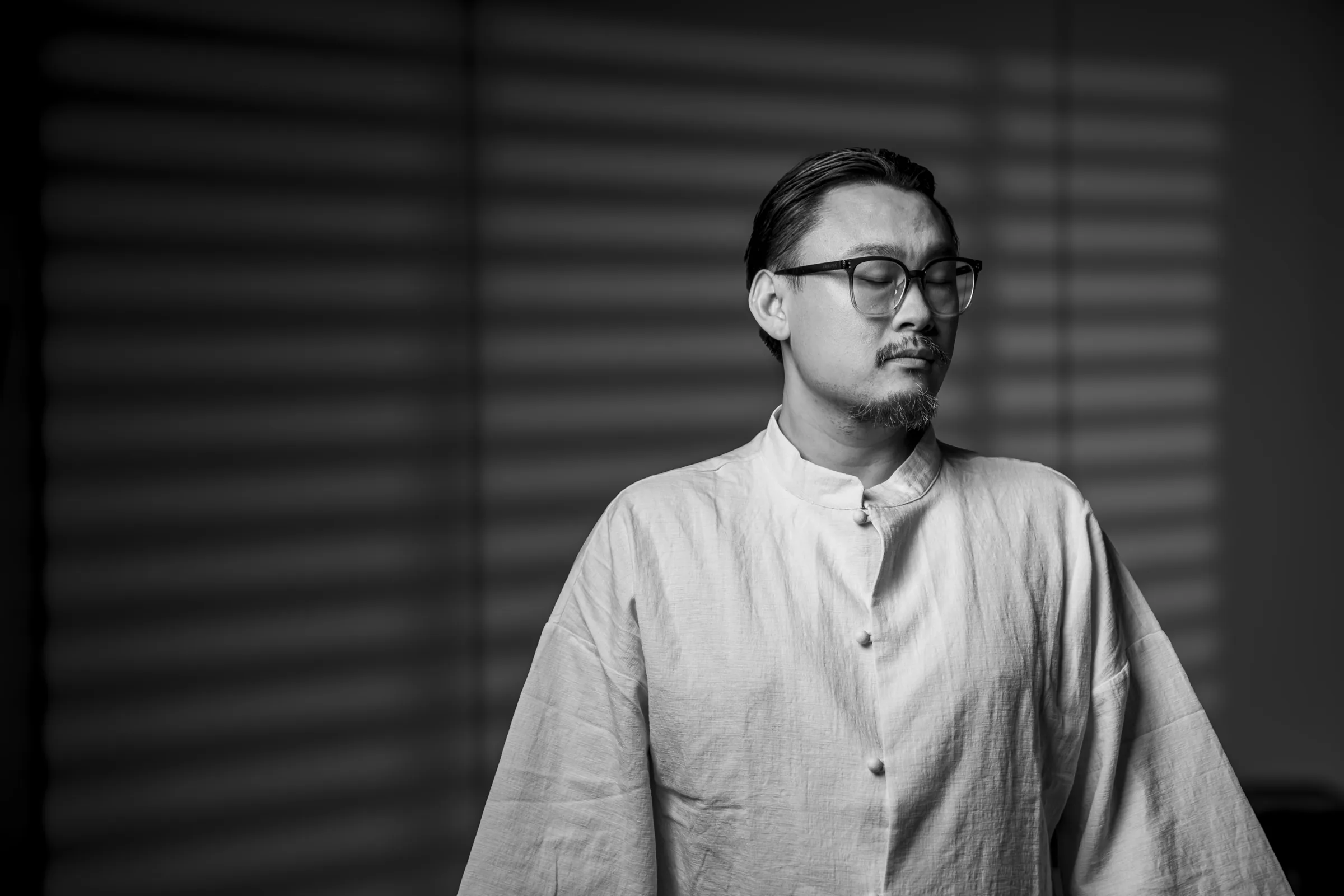 Master Lao standing still in slatted window light, eyes closed.
